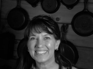 Bren smiling in front of her cast iron that is displayed behind her on the log walls of the homestead house.